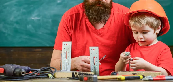 Pai como conceito de faz-tudo. Rapaz, criança de capacete protector faz à mão, reparar, faz artesanato com o pai. Pai, pai com barba e filho pequeno na sala de aula, quadro no fundo — Fotografia de Stock