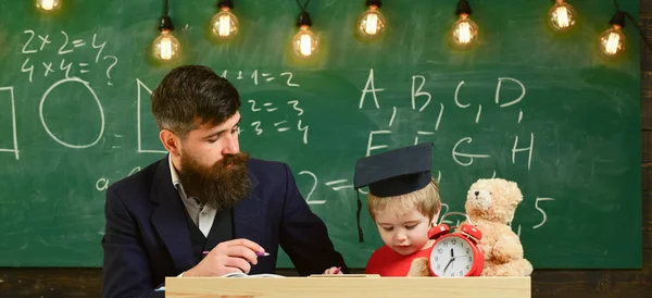 Fadern lär son matematik. Entusiastiska barn studerar med lärare. Matematik lektion koncept. Lärare i formella slitage och elev i mortarboard i klassrummet, svarta tavlan på bakgrund — Stockfoto