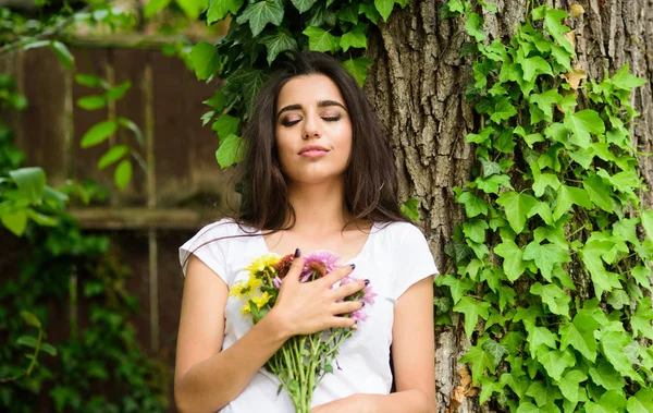 Girl gorgeous brunette hugs bouquet flowers while lean on tree trunk with ivy. Girl dreamy remembering great date. Woman enjoy bouquet flowers. That was great date. She got her favorite flowers — Stock Photo, Image