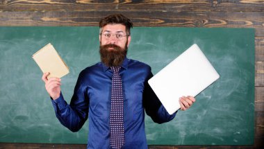 Teacher bearded hipster holds book and laptop. Choose right teaching method. Modern instead outdated. Teacher choosing modern teaching approach. Modern technologies benefit. Digital against paper
