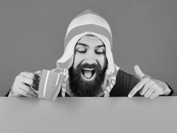 Ideia de bebida de Outubro. Homem de chapéu quente mantém copo azul no fundo verde e amarelo, espaço cópia. Hipster com barba e sorriso aponta para baixo e mantém o chá — Fotografia de Stock