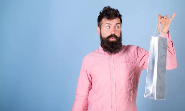 Hipster on excited face shopping addicted or shopaholic. Guy shopping on sales season with discounts. Sale and discount concept. Man with beard look at shopping bag, light blue background, copy space — Stock Photo, Image