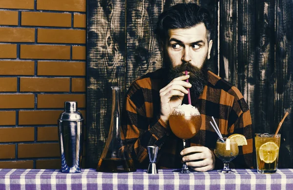 Barman z brodą i zaskoczony twarz pije ze szkła ze słomką koktajl. Człowiek pije koktajl na podłoże drewniane. Koncepcja alkoholu i koktajle. — Zdjęcie stockowe