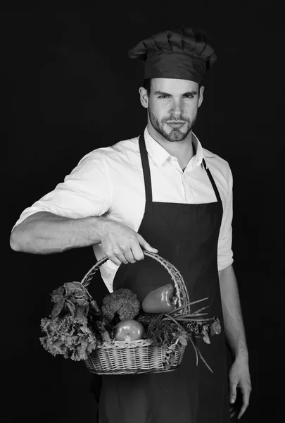 Kock i vinrött förkläde och hatt innehar sallad, paprika, tomat, gurka och chili. Hälsosam matkoncept. Mannen med skägg på svart bakgrund. — Stockfoto