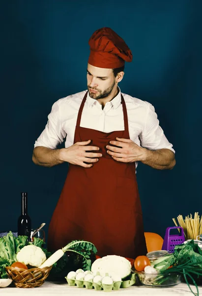 Man in burgundy cook hat and apron. Cook works in kitchen near table with vegetables and tools.