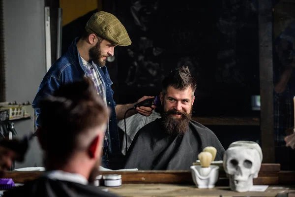 Barber and hipster client with beard checking haircut in mirror, dark background. Hipster and barber talking during haircut session. Service concept. Man with beard explain hairstyle he prefers