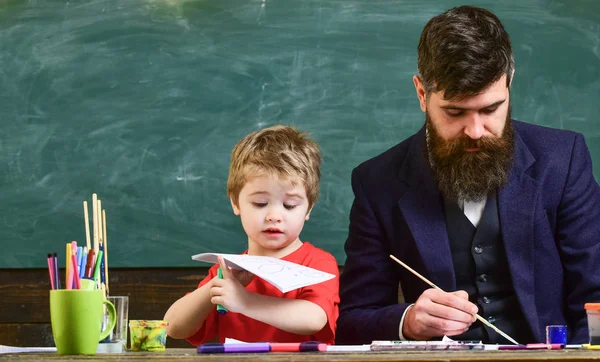 Teacher with beard, father and little son in classroom while drawing, creating, chalkboard on background. Child and teacher on busy faces painting, drawing. Arts lesson concept