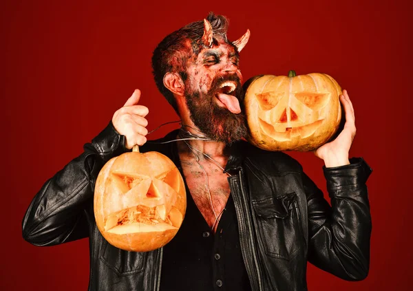 Demon with horns and cheeky face licks carved jack o lanterns. Devil or monster with October decorations. Man wearing scary makeup — Stock Photo, Image