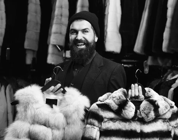 El tipo con la cara feliz muestra abrigos de piel en la tienda de moda. Asistente de tienda sostiene gris sable y rayas abrigos de piel de chinchilla . — Foto de Stock