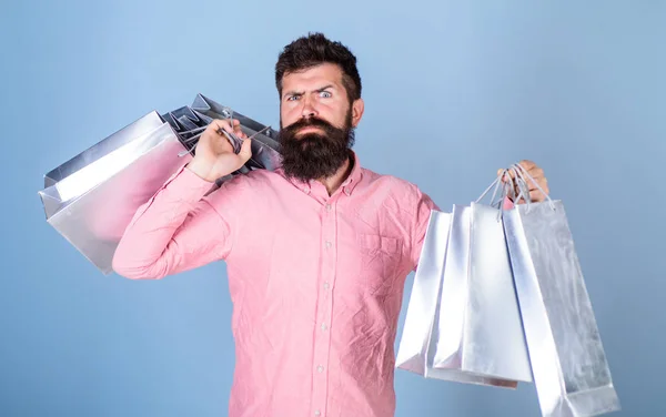Sale and discount concept. Guy shopping on sales season with discounts. Man with beard and mustache carry shopping bags, light blue background. Hipster on strict face shopping addicted or shopaholic
