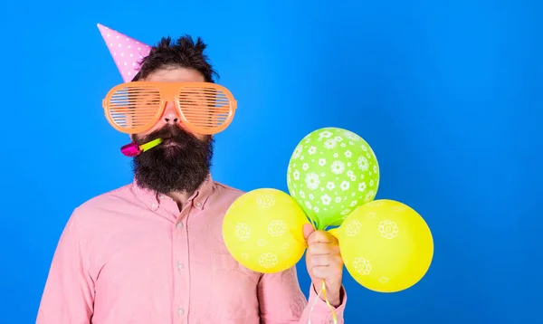 Celebration koncept. Hipster i giant solglasögon firar födelsedag. Killen i partiet hatt med air ballonger firar. Mannen med skägg och mustasch på lugna ansikte blåser in part horn, blå bakgrund — Stockfoto