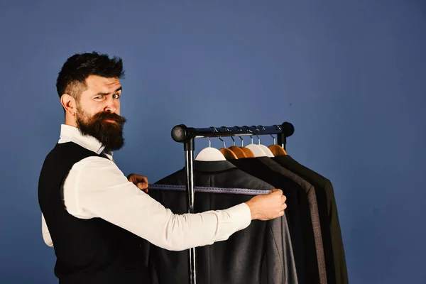 Businessman with confused face near jackets on blue background. Man with beard in vest by clothes rack. Fashion choice