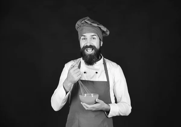 Hombre o hipster con barba sostiene utensilios de cocina sobre fondo negro. Cocinar con la cara feliz en uniforme de color burdeos sostiene tazón, batir —  Fotos de Stock