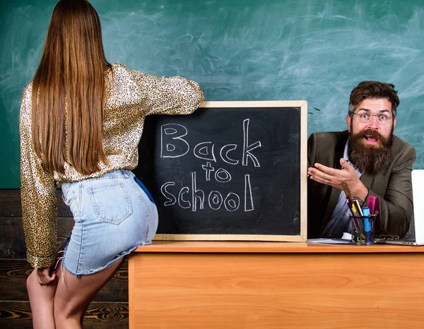Teacher indignant sit table chalkboard background. School discipline and behaviour rules. Student girl seduces experienced teacher. Student in mini skirt with nice buttocks stand near blackboard