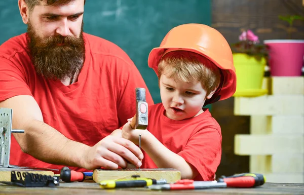 Küçük Yardımcısı kavramı. Babası öğretim araçları, çekiç, kullanmak için küçük oğlu sakal kara tahta arka plan ile. Çocuk, çocuk ile baba çekiç kullanmayı öğrenme koruyucu kask içinde meşgul