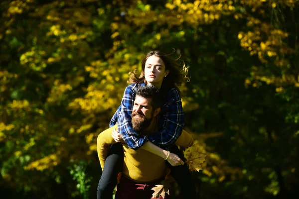 Man and woman with happy faces on natural background. Girl and bearded guy or happy lovers have fun outdoors having piggyback ride. Couple in love walks in autumn park. Freedom and fall season concept