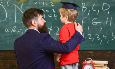 Child in graduate cap listening teacher, chalkboard on background, rear view. Teacher with beard, father teaches little son in classroom, chalkboard on background. Instructive conversation concept clipart