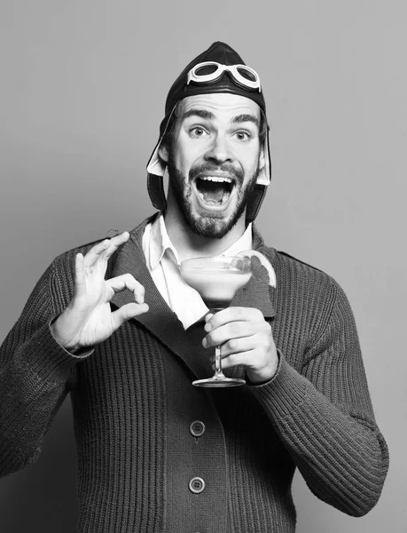Coquetel alcoólico em suéter de malha cinza com chapéu e óculos no fundo de estúdio vermelho. piloto barbudo com coquetel em vidro — Fotografia de Stock