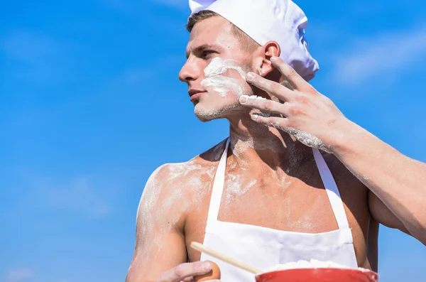 Concepto Baker. Cocine o chef con hombros musculares sexy y pecho cubierto de harina. Cocinero cocinar la preparación de la masa para hornear con harina. Hombre en la cara ocupada lleva sombrero de cocina y delantal, cielo en el fondo — Foto de Stock