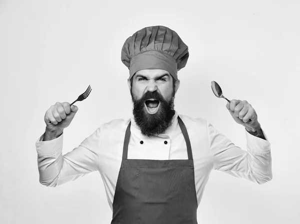 Homme à la barbe tient fourchette et cuillère sur fond blanc. Cuisiner avec le visage en colère en uniforme bordeaux montre couverts — Photo
