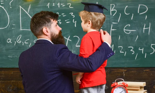 Child in graduate cap listening teacher, chalkboard on background, rear view. Teacher with beard, father teaches little son in classroom, chalkboard on background. Instructive conversation concept — Stock Photo, Image