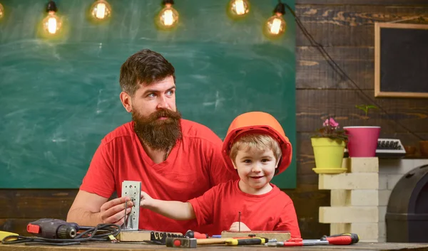 Father with beard teaching little son to use tools in classroom, chalkboard on background. Boy, child busy in protective helmet makes by hand, repairing with dad. Little assistant concept
