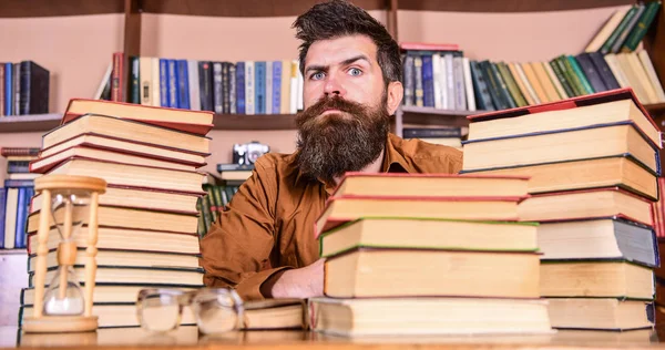 Homem no rosto estrito sentar entre pilhas de livros, enquanto estuda na biblioteca, estantes de livros no fundo. Professor ou estudante com barba sentar-se à mesa com ampulheta e óculos, desfocado. Conceito de bicho de biblioteca — Fotografia de Stock