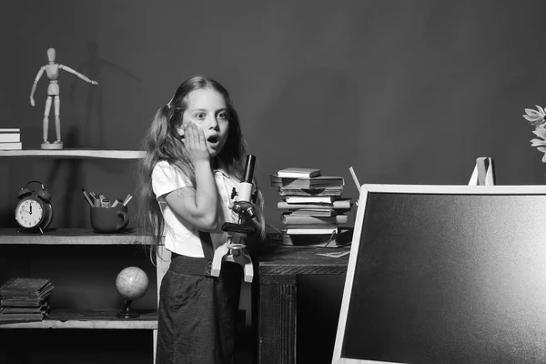 Schoolgirl with shocked face in her classroom. Girl holds microscope and stands by blackboard, copy space — Stock Photo, Image