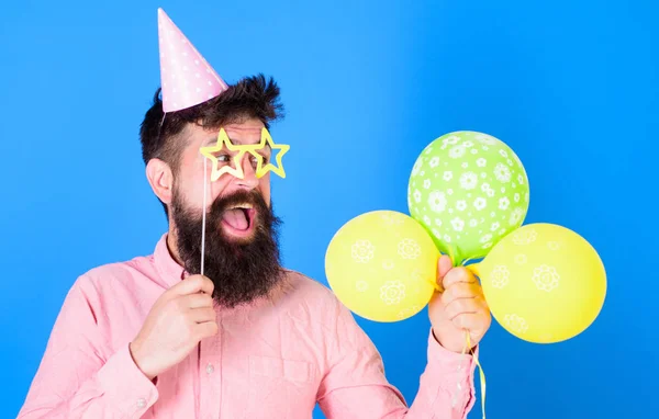 Homem com barba e bigode no rosto feliz segurar balões de ar, fundo azul. O tipo de chapéu de festa com atributos de férias celebra. Hipster em óculos em forma de estrela celebra o aniversário. Conceito de aniversário — Fotografia de Stock