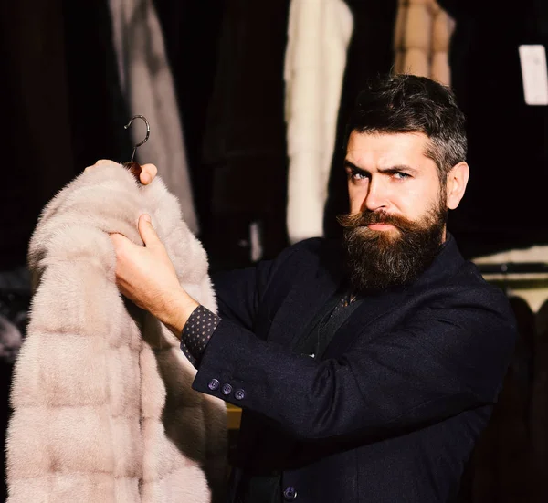 Macho con aspecto elegante con piel de visón en la tienda de moda. Hombre con barba y bigote sostiene abrigo de piel . — Foto de Stock