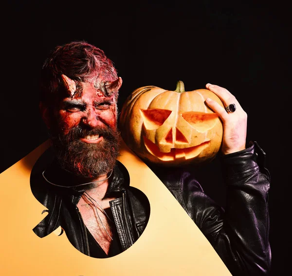 Demon with horns and evil smile holds jack o lantern. Halloween party concept. Man wearing scary makeup holds carved pumpkin