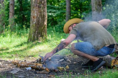 Şenlik için Ultimate guide. Ormanda twigs veya ahşap sopa bir piramit gibi duran ve yaprakları altında yer. Adam hasır şapka ormandaki şenlik ateşi hazırlar. Nasıl açık havada şenlik ateşi kurmak için