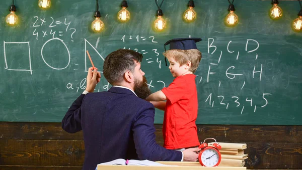 Ung manlig lärare förklarar ämne, vägleder sitt barn student till lärande medan sitter i klassrummet, svarta tavlan med klotter på bakgrund, bakifrån — Stockfoto