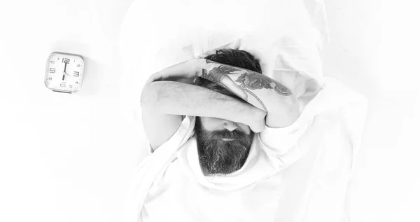 Tattooed hipster in morning, near alarm clock, white background. Young man relaxation on the bed top view, hiding face.