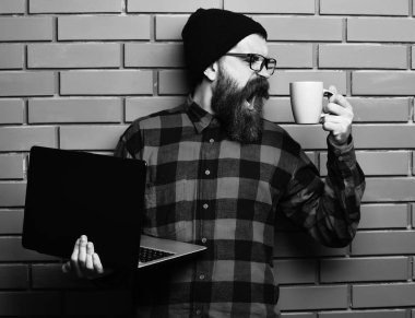 Laptop mag veya Kupası ile şapka ve gözlük kahverengi tuğla duvar studio arka plan üzerinde kırmızı siyah damalı gömlek holding hippi. Sakallı acımasız beyaz hipster laptop Kupası ile tutarak