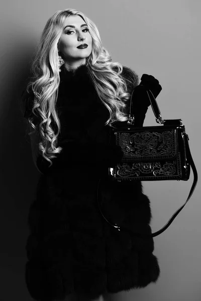 young fashionable sexy pretty woman with beautiful long curly blonde hair in waist coat of burgundy fur and black velvet gloves showing white fashion bag on red studio background