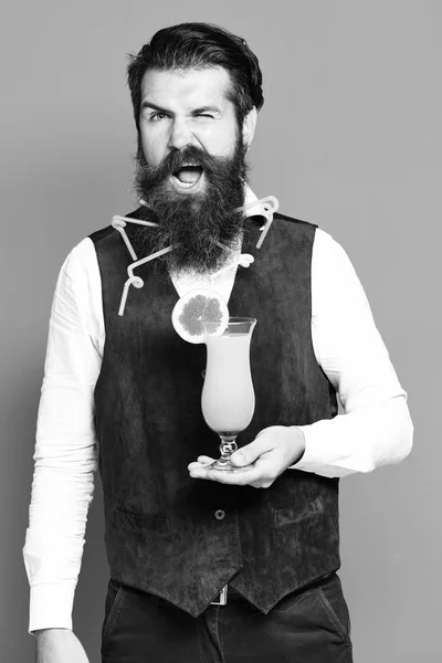 Handsome bearded man with colorful tubes in long beard and mustache has stylish hair on funny face holding glass of alcoholic cocktail in vintage suede leather waistcoat on red studio background — Stock Photo, Image