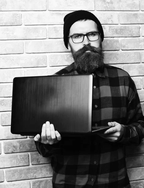 Hipster segurando laptop em vermelho camisa xadrez preto com chapéu e óculos no fundo do estúdio parede de tijolo bege. Barbudo brutal caucasiano hipster segurando laptop — Fotografia de Stock