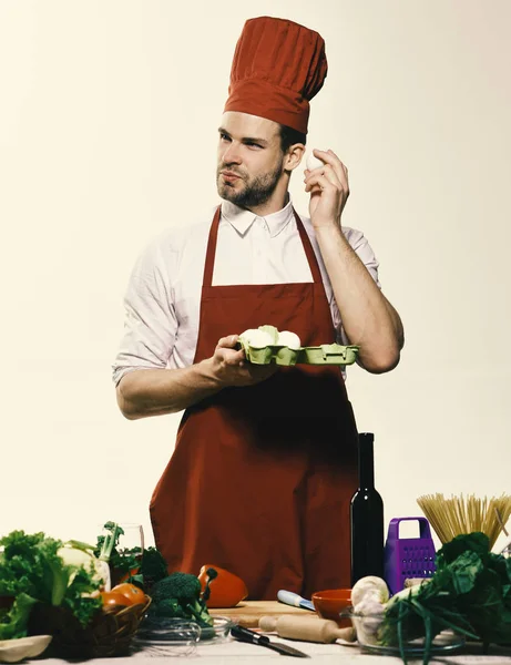 Cook står vid bordet med köksutrustning och grönsaker. Kock i Bourgogne enhetliga håller ägg. Köksutrustning och mat koncept. — Stockfoto