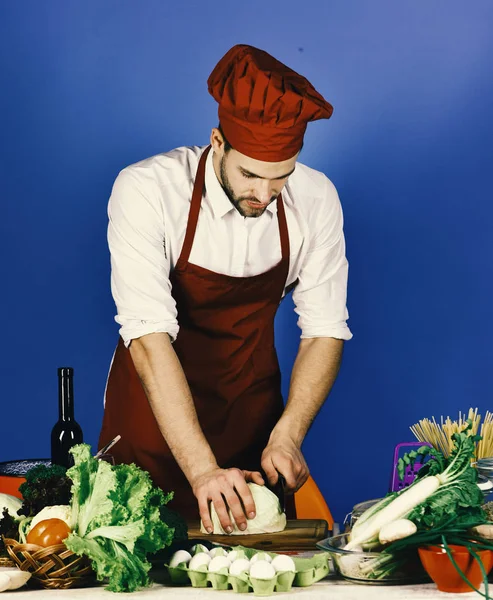 Cook fungerar i köket nära bord med grönsaker och verktyg. Kock med upptagen ansikte kotletter kål med kniv på blå — Stockfoto