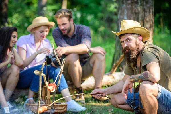 Hipster roasting sausage while friends sit on log sharing impression and watching photos on camera. Tourists having snack time with roasted over fire food. Friends group tourist relaxing near bonfire