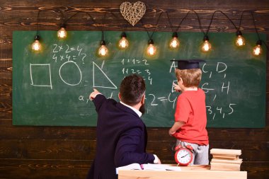 Teacher with beard, father teaches little son in classroom, chalkboard on background. Basics of education concept. Boy, child in graduate cap looks at scribbles on chalkboard while teacher explains clipart