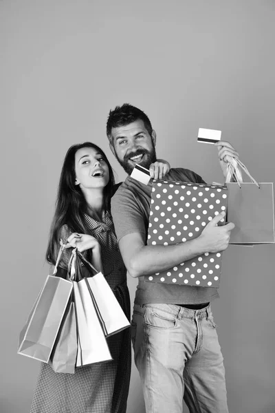 Couple in love holds shopping bags on pink background. Man with beard holds credit card and polka dotted box. Shopping and leisure concept. Guy with beard and girl