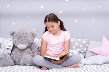 Uyku önce okuyun. Kız çocuk kitap okumak bir oyuncak ayı ile yatağa otur. Çocuk yatmaya hazırlanın. Keyifli zaman rahat yatak odasında. Kız çocuk uzun saçlı şirin pijama rahatla ve oyuncak ayı için kitap okumak.