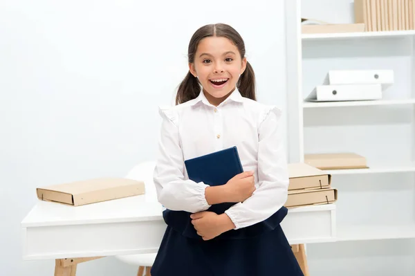 Рад вступить в первый класс. Девочка держит книжный столик на белом фоне. Школьнице нравится учиться и посещать уроки. Детская школьная форма улыбающееся лицо любит посещать школьные уроки — стоковое фото