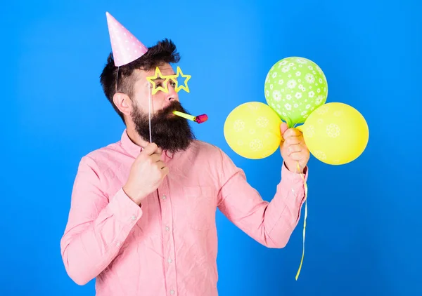 Mann mit Bart im Gesicht hält Luftballons, blauer Hintergrund. Hipster in sternförmiger Brille bläst ins Partyhorn. Überraschungskonzept. Mann im Partyhut mit Urlaubsattribute feiert — Stockfoto