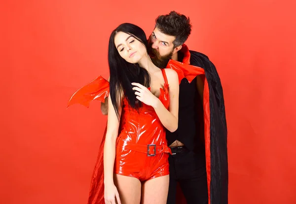 Couple in love play role game. Vampires victim concept. Man and woman dressed like vampire, demon, red background. Vampire in cloak behind sexy devil girl. Vampire bites female neck