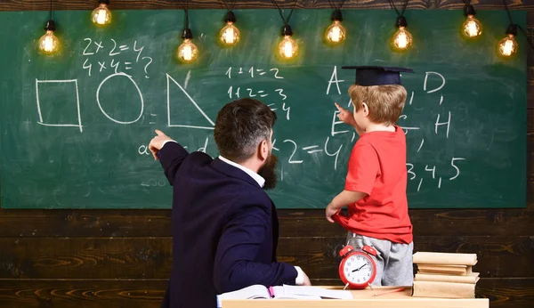 老师带着胡子, 父亲在教室里教小儿子, 黑板上的背景。男孩, 研究生帽的孩子看黑板上的涂鸦, 而老师解释。教育理念基础 — 图库照片