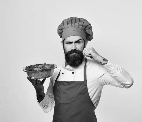 Koken met ernstige gezicht Bourgondië uniforme houdt gebakken schotel. Man met baard geïsoleerd op witte achtergrond. — Stockfoto