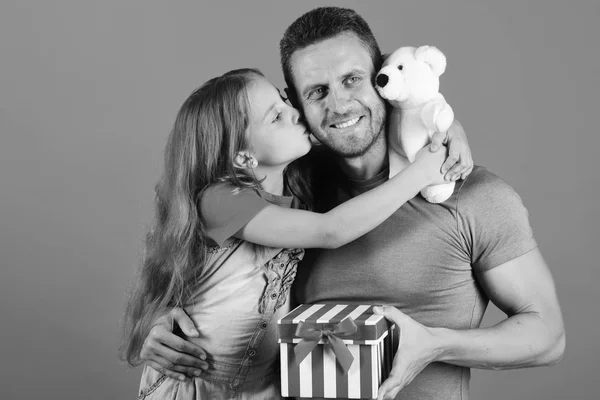 Girl and man with happy faces on red background. Kid kisses her daddy. Schoolgirl and dad hold present box and white teddy bear.